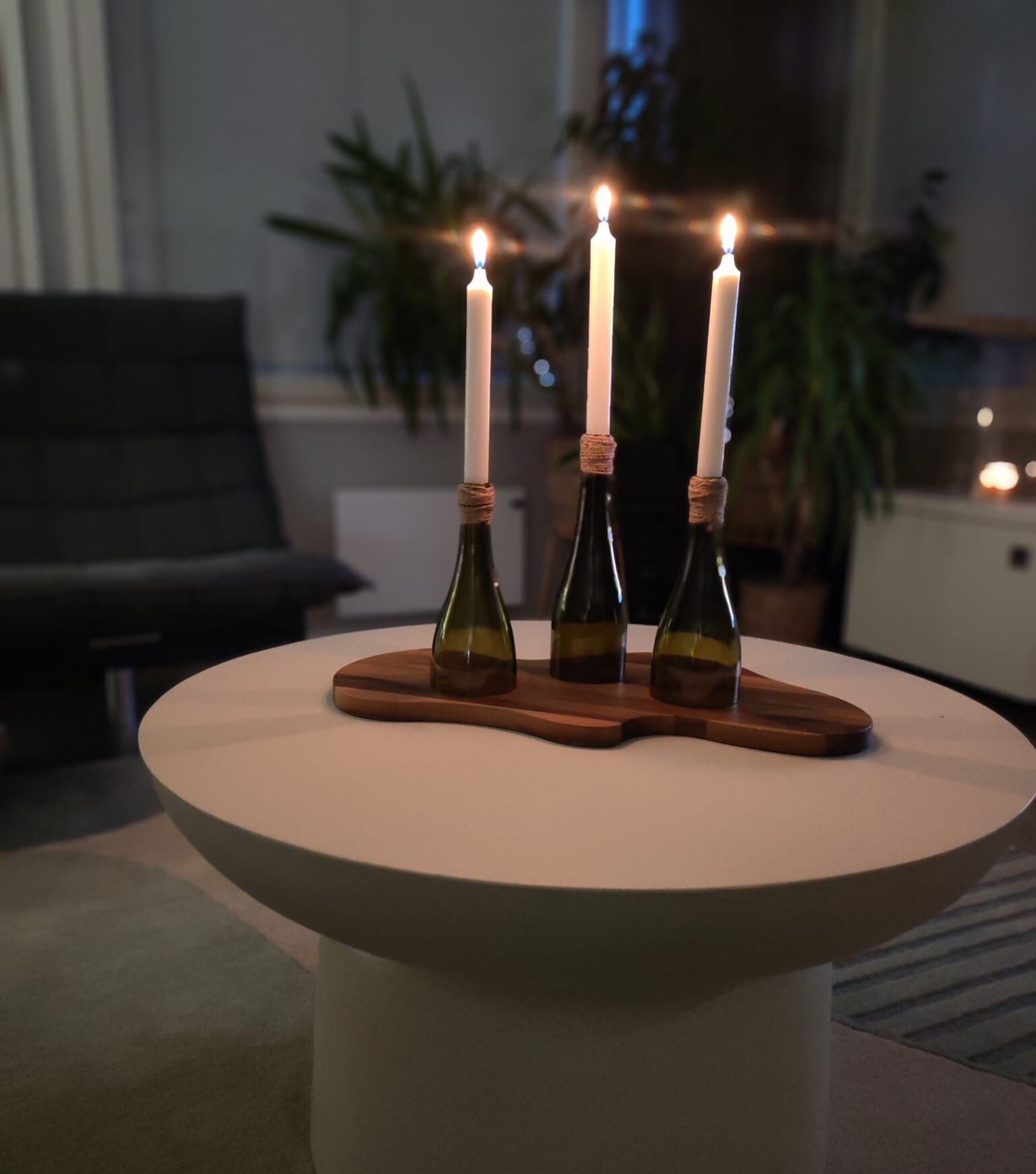 Kolme pullonkaulasta tehtyä kynttilänjalkaa palavine kruunukynttilöineen puualustalla olohuoneen pöydällä illalla.