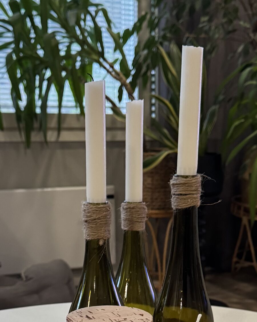 Kolme pullonkaulasta tehtyä kynttilänjalkaa, kaulat kiedottu narulla, kruunukynttilät paikallaan.