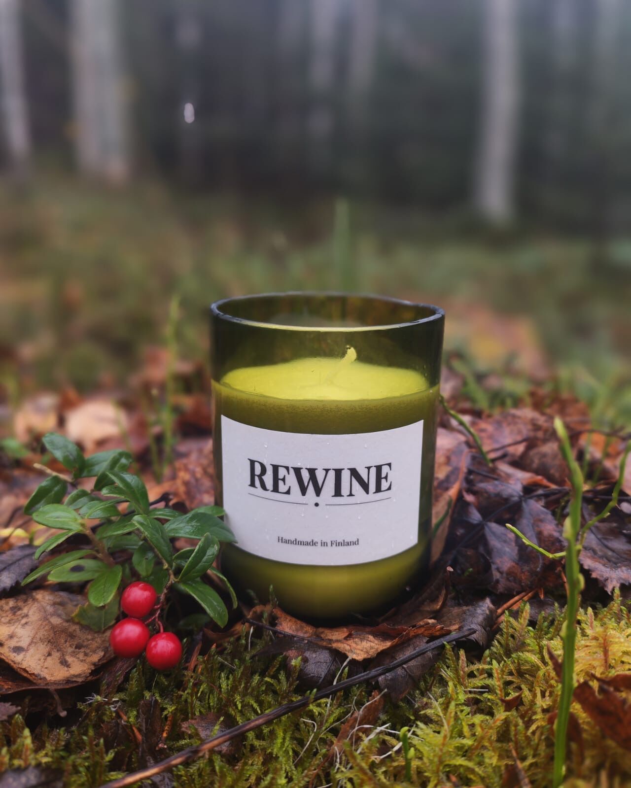 REWINE-kynttilä leikatussa vihreässä viinipullossa syksyisellä metsänpohjalla puolukoiden vieressä.